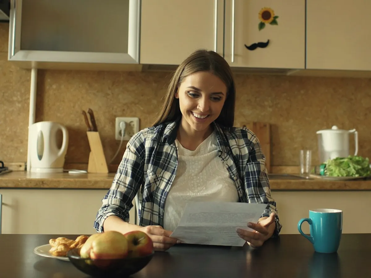 A woman reads a letter at her kitchen table with coffee and fruit.